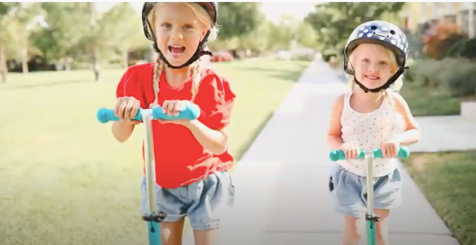 Two kids in helmets riding Micro Kickboard scooters down a sidewalk