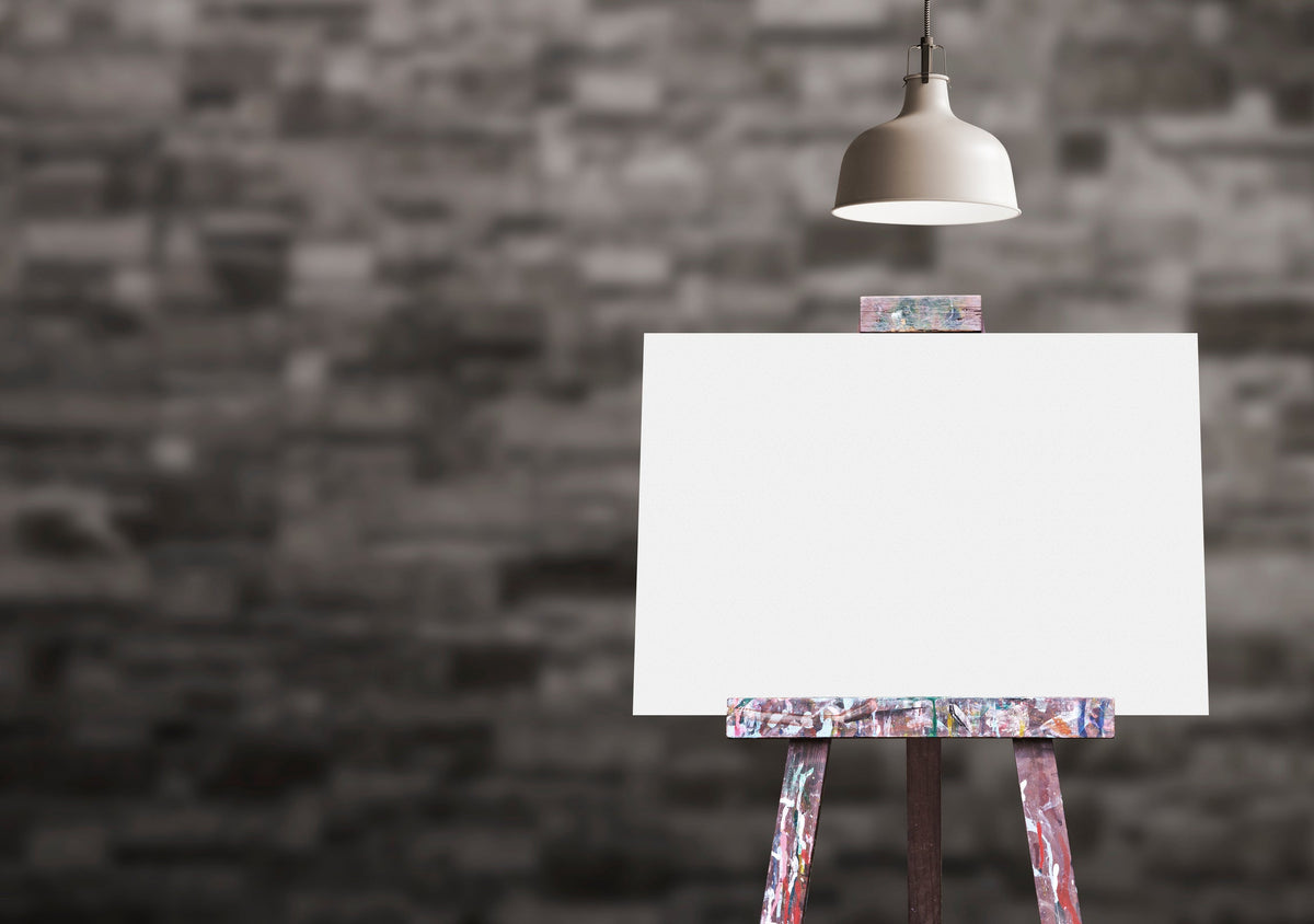 A blank canvas on a wooden easel under a single spotlight against a dark brick wall
