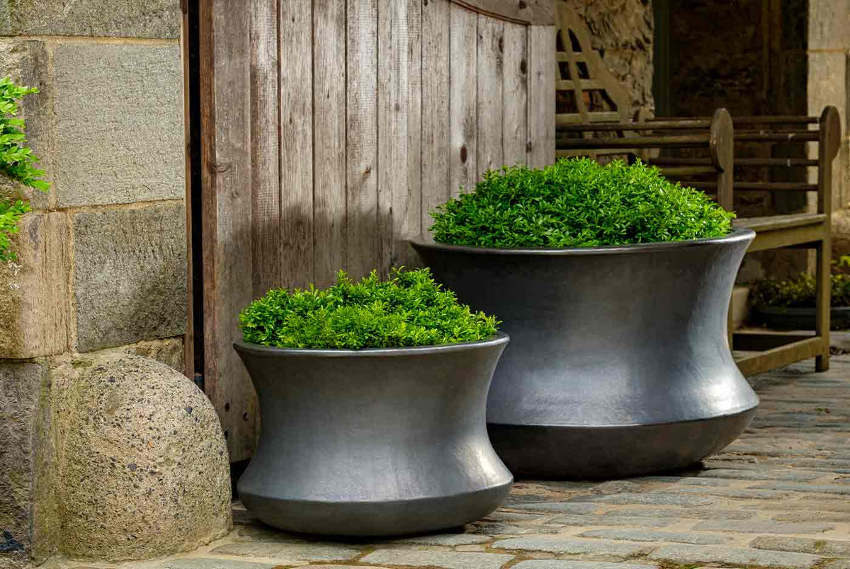 Two dark gray Campania planters filled with boxwood on a cobblestone courtyard