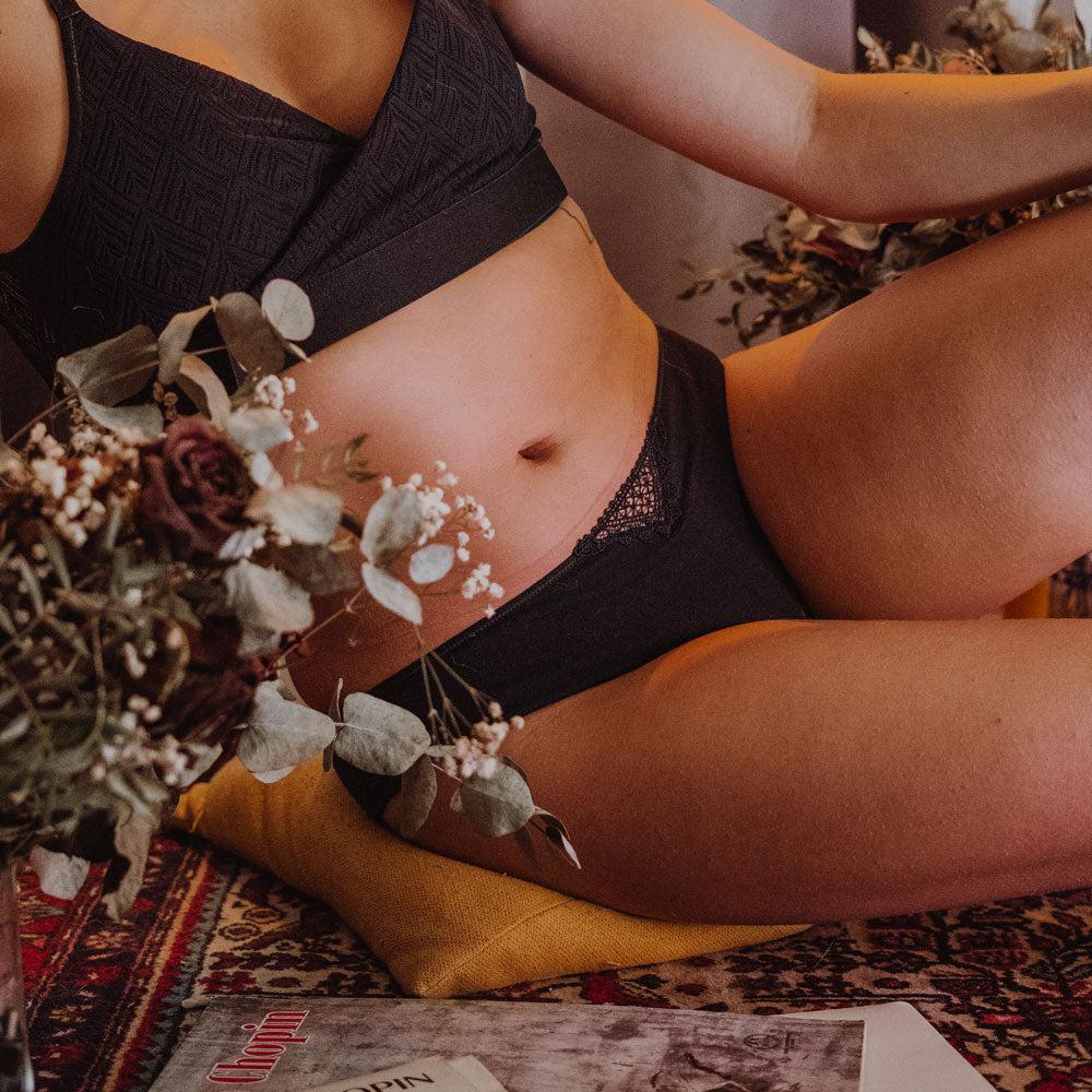 Model wearing black Elia lingerie, lying on a mustard cushion beside dried flowers